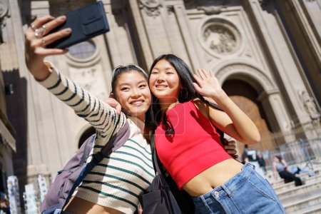 Téléchargez les photos : Joyeux jeunes femmes asiatiques touristes en tenues décontractées souriant et prenant autoportrait sur smartphone tout en se tenant contre la cathédrale de Grenade en Espagne pendant les vacances - en image libre de droit