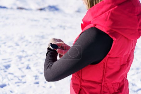 Téléchargez les photos : Vue latérale d'une jeune femelle anonyme vêtue de vêtements chauds tenant une montre-bracelet et vérifiant l'heure lorsqu'elle se tient debout sur une pente enneigée floue en plein jour en hiver - en image libre de droit
