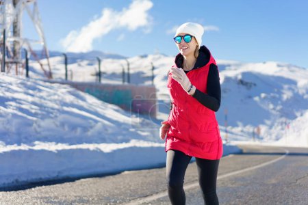 Téléchargez les photos : Jeune femme souriante en gilet rouge bonnet bonnet lunettes de soleil polarisées gants regardant loin, tout en faisant du jogging sur la route asphaltée près de la neige contre la montagne enneigée et le ciel bleu en plein jour - en image libre de droit