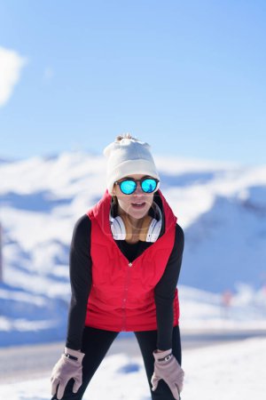 Foto de Joven mujer sana en ropa de abrigo y gafas de sol descansando mientras se apoya sobre las rodillas contra las montañas en la nieve - Imagen libre de derechos