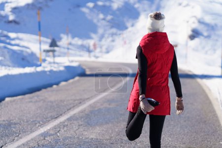Foto de Vista posterior de la mujer anónima en ropa deportiva caliente calentando la pierna en la carretera vacía en la naturaleza nevada a la luz del sol - Imagen libre de derechos