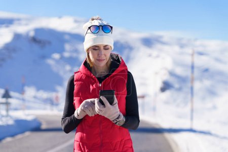 Téléchargez les photos : Femme athlétique adulte en vêtements chauds smartphone navigation tout en se tenant debout sur la route asphaltée dans les montagnes d'hiver - en image libre de droit