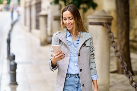 Foto de Mujer adulta feliz en ropa casual y abrigo gris elegante sonriendo y viendo video en el teléfono inteligente mientras está de pie en el fondo borroso de la calle en la ciudad - Imagen libre de derechos