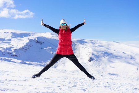 Photo for Full body of excited young female in warm clothes cap, polarized sunglasses looking at camera while jumping in air with widened legs stretched arms against snowy mountains and blue sky - Royalty Free Image
