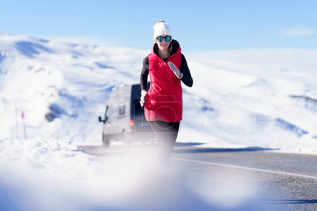 Photo for Smiling female wearing warm vest and hat with sunglasses jogging on road in amazing mountains in winter - Royalty Free Image