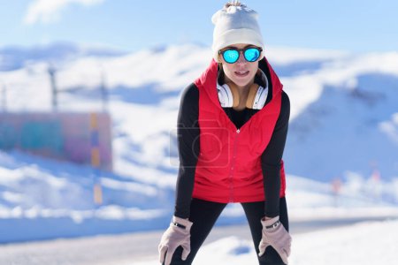 Téléchargez les photos : Femme sportive calme dans des vêtements chauds et des lunettes de soleil appuyé sur les genoux récupérer respirer tout en faisant du jogging dans la nature - en image libre de droit