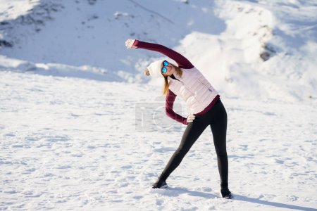 Photo for Full body of young female in cap polarized sunglasses, looking away and stretching arm sideways while standing with legs apart and hand on thigh on snowy mountain in daylight - Royalty Free Image