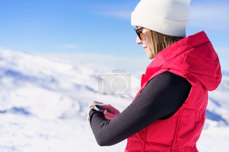 Téléchargez les photos : Vue latérale de la jeune femme vêtue de lunettes de soleil chaudes, tenant la montre-bracelet et regardant vers le bas pour vérifier l'heure tout en se tenant debout sur une pente de montagne enneigée floue en plein jour - en image libre de droit