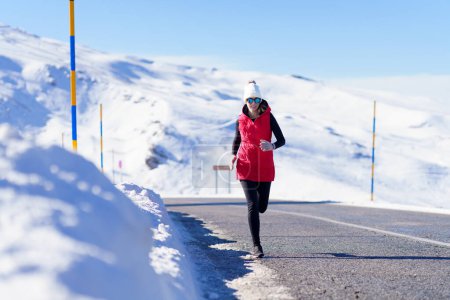 Foto de Cuerpo completo de mujer en ropa de abrigo y gafas de sol trotando en el camino de asfalto con montañas nevadas - Imagen libre de derechos