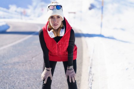 Téléchargez les photos : Femme sportive déterminée en vêtements chauds et avec écouteurs appuyés sur les genoux tout en regardant la caméra sur la route en hiver - en image libre de droit