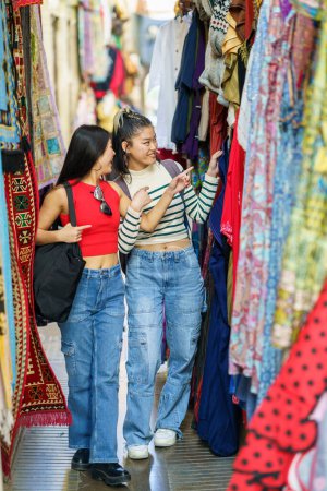 Foto de Chicas sonrientes amigas en ropa casual caminando a lo largo de colorida tienda de ropa en el mercado callejero de la ciudad durante las vacaciones de verano - Imagen libre de derechos