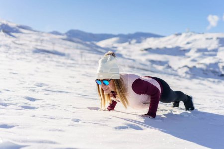 Photo for Cheerful young female in warm clothes and sunglasses lying in Chaturanga pose on white snowy slope of mountain on sunny winter day with blue sky - Royalty Free Image