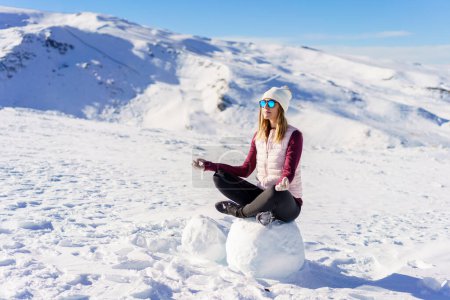 Téléchargez les photos : Corps complet de la jeune voyageuse en vêtements décontractés et lunettes de soleil assis sur la neige dans la pose du lotus et regardant loin sur la journée ensoleillée d'hiver - en image libre de droit