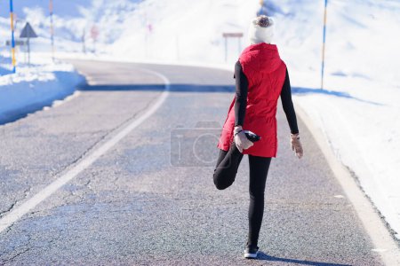 Foto de Vista posterior de la señora atlética anónima en ropa de abrigo que estira las piernas mientras está de pie en el camino de asfalto en las montañas nevadas - Imagen libre de derechos