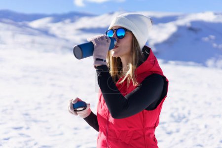 Photo for Side view of young female in warm clothes, beanie wool cap polarized sunglasses drinking warm water from thermos bottle and looking away while standing on snowy mountain slope in daylight - Royalty Free Image