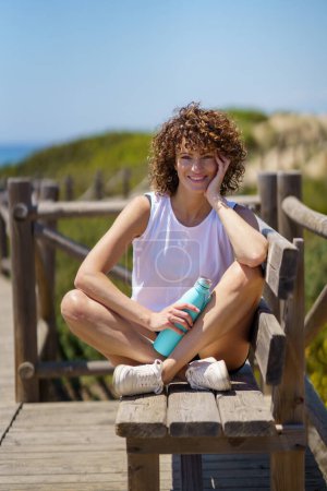 Foto de Joven atleta en ropa deportiva sonriendo y mirando a la cámara mientras está sentada en un muelle de madera con una botella de agua sobre un fondo borroso - Imagen libre de derechos