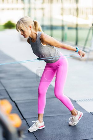 Téléchargez les photos : Vue latérale de l'athlète féminine adulte en forme dans des leggings roses regardant loin tout en se tenant debout sur la pointe des pieds et l'exercice, avec bande de résistance pendant l'entraînement de remise en forme dans la salle de gym en plein jour - en image libre de droit