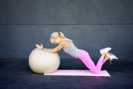 Photo for Side view of adult female in sportswear, ponytail looking down while lying with stretched hands on fit ball on gym floor and knees of folded up legs on mat in daylight - Royalty Free Image