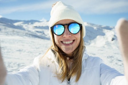 Téléchargez les photos : Jeune femme positive en bonnet de bonnet de vêtements chauds, et lunettes de soleil polarisées souriant et regardant la caméra tout en prenant selfie et debout sur la pente de montagne enneigée par une journée ensoleillée en hiver - en image libre de droit