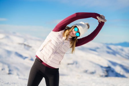 Téléchargez les photos : Jeune femme positive en vêtements de sport et lunettes de soleil étirant les bras latéralement dans la pose de yoga et regardant la caméra tout en se tenant debout sur le fond des montagnes enneigées - en image libre de droit