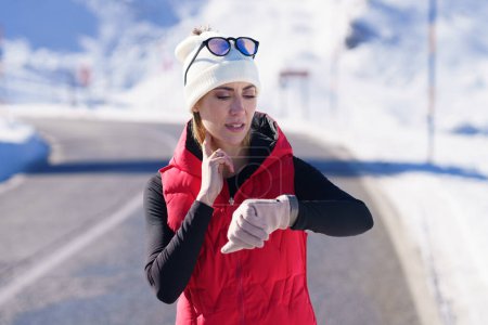 Téléchargez les photos : Femme sportive adulte en vêtements chauds synchronisation battement de coeur tenant les doigts sur le cou tout en vérifiant le pouls pendant l'entraînement cardio en hiver nature - en image libre de droit
