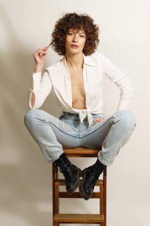 Photo for Full length of flirty female model with curly hair, in stylish wear resting on wooden stool playing with curl and looking at camera against white background - Royalty Free Image
