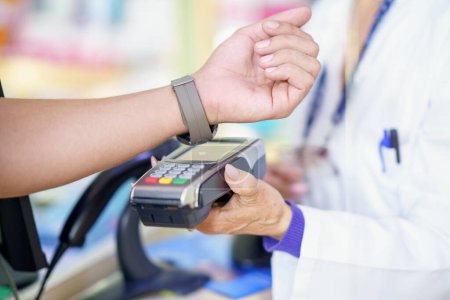 Téléchargez les photos : Homme méconnaissable payant par smartwatch avec dataphone dans une pharmacie, assisté par une pharmacienne détenant le système de paiement. - en image libre de droit