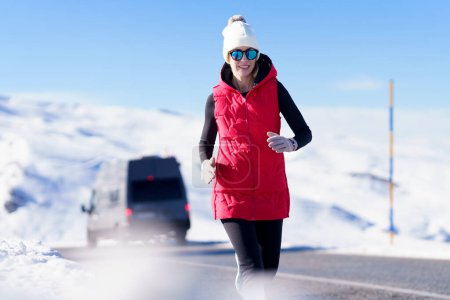Téléchargez les photos : Femme sportive souriante en vêtements chauds et lunettes de soleil courant sur la chaussée avec des montagnes enneigées par une journée ensoleillée - en image libre de droit
