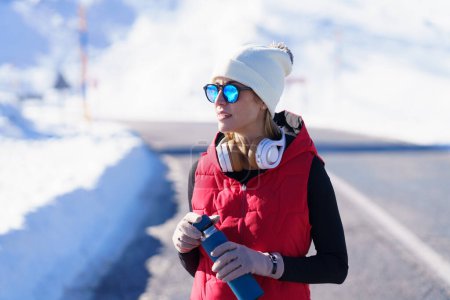 Téléchargez les photos : Jeune femme inspirée en vêtements chauds et lunettes de soleil tenant thermos bouteille tout en ayant l'entraînement dans les montagnes enneigées - en image libre de droit