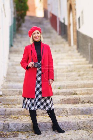 Photo for Full body of self assured young blondie, with photo camera on neck in stylish red coat and beret smiling happily while standing on stone steps between aged buildings in town - Royalty Free Image