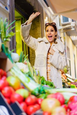 Téléchargez les photos : Angle bas de la jeune femme positive en vêtements chauds, souriant et regardant la caméra tout en agitant la main et debout près de décrochage avec divers légumes et fruits en plein jour - en image libre de droit