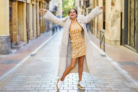 Photo for Stylish young woman wearing fashionable long coat with dress and shoe dancing on street against building background - Royalty Free Image