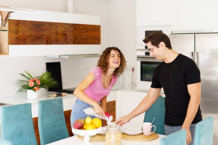 Photo for Positive couple having fun while standing near dining table laughing female with eyes closed, pouring juice and joking looking down male in eyeglasses holding glass in kitchen - Royalty Free Image