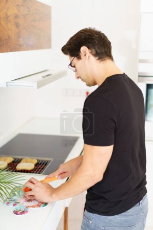 Photo for Side view of adult male in casual clothes and eyeglasses looking down while standing in kitchen near cooking range, with bread slices on grill and cutting tomato with knife in daylight - Royalty Free Image