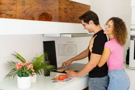 Photo for Side view of happy adult couple while standing near cooking range smiling looking down male in eyeglasses toasting bread slices, on grill and female with eyes closed hugging from behind in daylight - Royalty Free Image