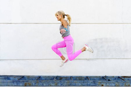 Téléchargez les photos : Vue latérale de la jeune femme en vêtements de sport sautant au-dessus du sol et souriant pendant l'entraînement dans la rue contre un mur blanc pendant l'entraînement de remise en forme - en image libre de droit
