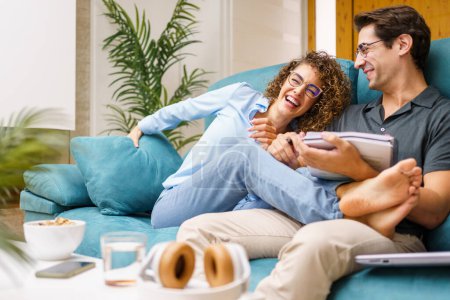 Photo for Cheerful young wife in casual clothes sitting with legs on husband, holding notebook while laughing and spending joyful moment together over couch in cozy living room - Royalty Free Image