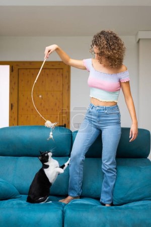 Foto de Joven mujer de pelo rizado en jeans y la parte superior de pie en el sofá y jugando con lindo gato en casa - Imagen libre de derechos