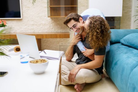 Téléchargez les photos : Aimant jeune femme embrassant l'homme par derrière tout en étant assis près d'un ordinateur portable et d'un ordinateur portable dans le salon à la maison - en image libre de droit