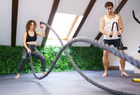Photo for Full body of young curly haired female athlete in sportswear exercising with battle ropes during workout with fitness buddy in modern gym - Royalty Free Image