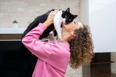 Photo for Side view of crop anonymous female in casual cloth with curly brown hair lifting and kissing adorable cat, with black and white fur while in room at home - Royalty Free Image