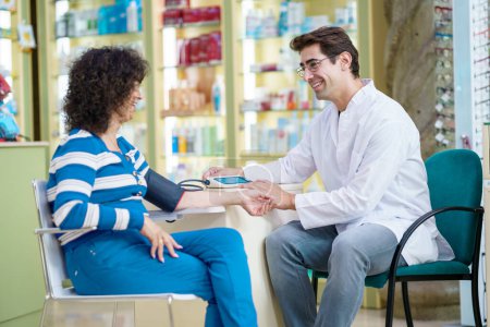 Téléchargez les photos : Vue latérale du pharmacien masculin heureux mesurant la pression artérielle de la femme assis avec le client et se regardant en pharmacie - en image libre de droit