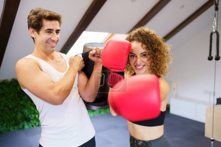 Photo for Smiling curly haired strong female fighter in red boxing gloves punching air during training with personal instructor holding bag in gym - Royalty Free Image