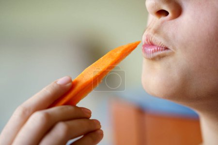 Téléchargez les photos : Cultiver anonyme jeune fille rondelle lèvres tout en tenant tranche de carotte fraîche à la maison - en image libre de droit