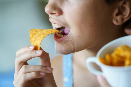 Photo for Crop anonymous teenage girl with mouth wide open about to eat yummy nacho chip at home - Royalty Free Image