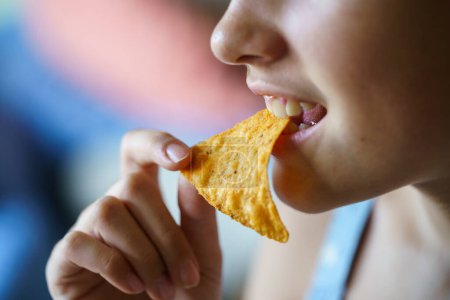 Photo for Closeup of crop unrecognizable happy teenage girl biting spicy nacho chip at home - Royalty Free Image