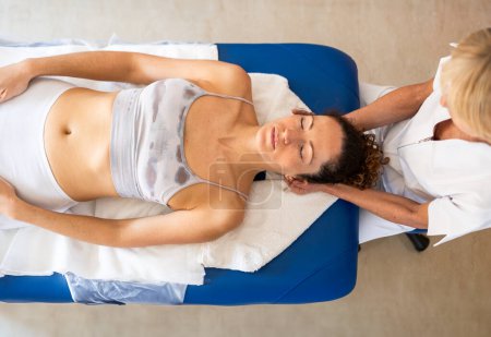 Téléchargez les photos : Vue du dessus de la tête de massage de physiothérapeute féminine pour la patiente couchée sur le canapé pendant la séance d'ostéopathie dans une clinique légère - en image libre de droit
