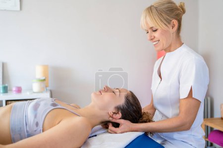 Photo for Side view of smiling female blond hair physiotherapist rubbing neck of client lying face up in bed and closing eyes during massage treatment - Royalty Free Image