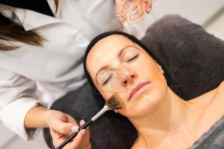 Téléchargez les photos : De dessus de la culture esthéticienne anonyme appliquer un produit cosmétique de soins de la peau avec brosse sur le visage de la femme dans le salon de beauté - en image libre de droit