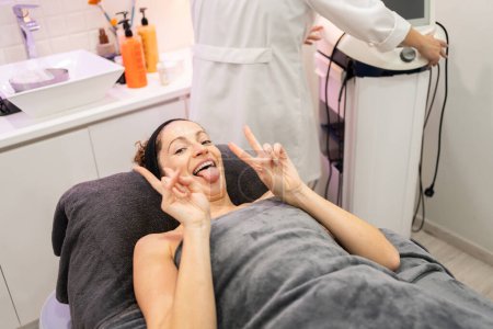 Photo for High angle of adult female with funny face and tongue out, looking at camera while lying on beauty salon bed with unrecognizable worker at equipment and showing V sign - Royalty Free Image